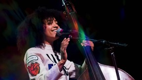 esperanza spalding, wearing a white jumpsuit with a "Life Force" logo, stands next to an upright bass