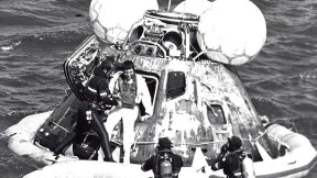 A man in a wetsuit helps an astronaut out of an Apollo capsule floating in the ocean