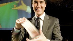 David Kwong shuffles the cards. Behind him, a video gives a closer look at his hands.