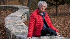 David Roberts seated outdoors on a stone wall by the Charles River