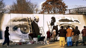Along a city street, mural on a concrete wall shows two men shaking hands, with a large pair of watchful eyes in  the background.