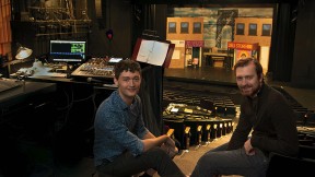 Finn Bamber and Clay Oxford sit beside a sound board with a theater set behind them