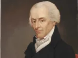 Portrait of a man with white hair, wearing a black coat, arms crossed, thoughtful expression.