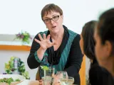 A woman in glasses gestures while speaking to two attentive listeners at a table.