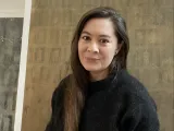 Woman with long hair, smiling, wearing a black sweater, in a textured beige background.