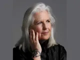 A woman with long, silver hair rests her chin on her hand, wearing a black top.