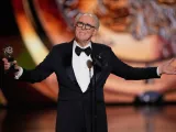 A man in a tuxedo gestures while holding an award on a stage.