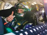 Racing driver gives a thumbs up from inside a car, wearing a helmet and safety gear.