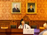 A man sitting at a wooden table in a formal setting with portraits on the wall.