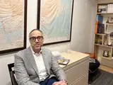 Man in a gray blazer sitting in an office with world maps on the wall and a bookshelf behind.