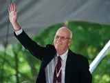 A man in a suit waves while smiling, with greenery in the background.