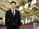 A man in a suit stands confidently on an ice rink, smiling, with a sports arena in the background.