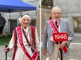 Older woman in a white outfit with a cane and red sash, alongside an elderly man in a gray suit holding a sign.
