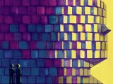 Two figures stand before a large, colorful pixelated face against a yellow background.