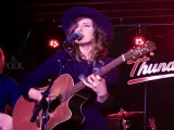 Singer performing on stage with a guitar, wearing a hat, and surrounded by band instruments.