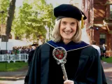 Katie O’Dair in academic regalia holds a ceremonial staff outdoors at a graduation ceremony.