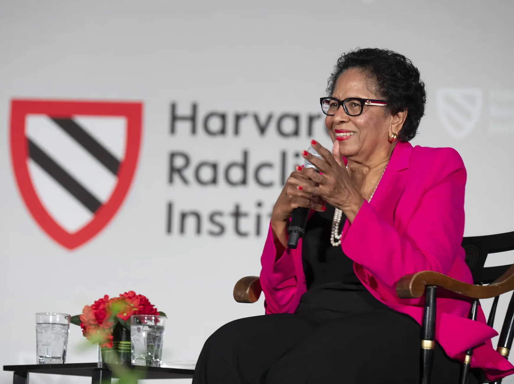 An African-American woman wearing a pink jacket sits in a chair and claps.