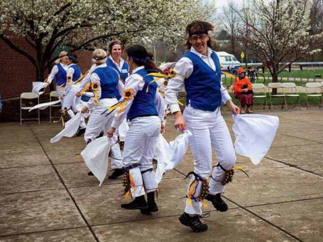 Morris dancers in white trousers and colorful vests wave handkerchiefs as they dance.