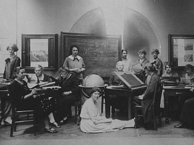 Several Harvard Computers and Radcliffe students pose for a photograph together, seated at desks and sitting on the floor