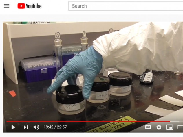 A human arm wearing a white goat and rubber glove in a lab setting handles three containers of lab chemicals