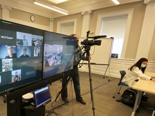 A hybrid classroom showing the large screens enabling a teacher to see all the students, remote or present, as he is followed by a camera operator.