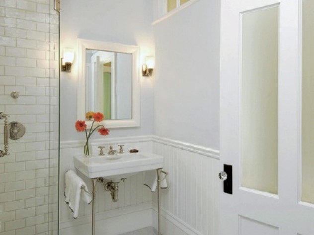 In an interior bathroom, transom windows and frosted glass allow natural light from an exterior window in the next room to reach the space. Allen chose to use period details such as wainscoting and molding to draw attention away from modern anachronisms like the frameless glass shower door. 