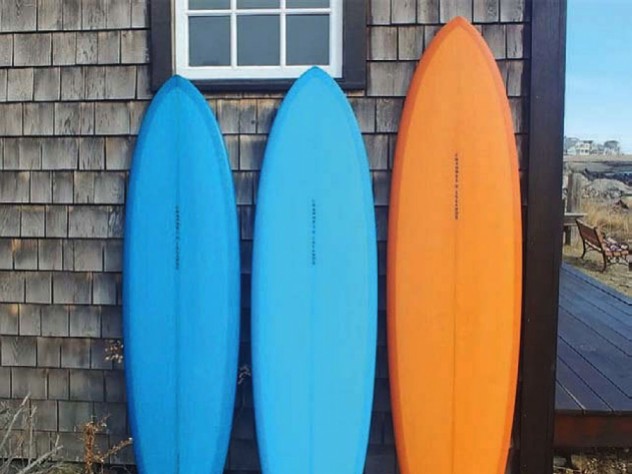 Two blue surfboards and an orange on rest on end against a weatherbeaten wall.