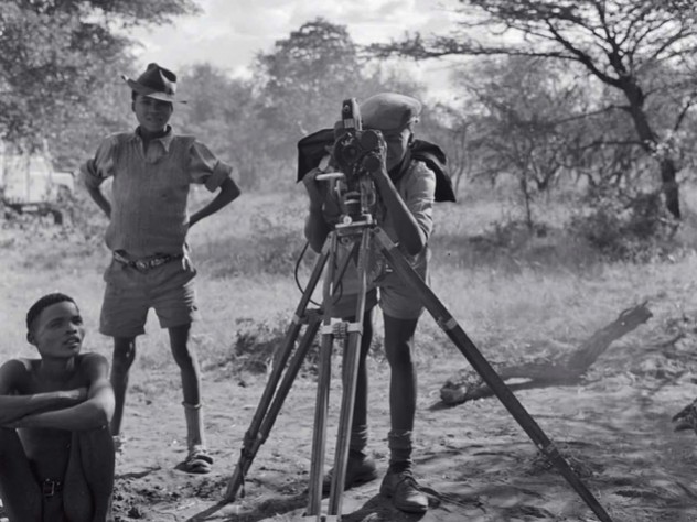 “/Gaishay focusing the camera,” a photograph taken during a 1957-58 ethnographic expedition in Namibia, shows three indigenous boys, one standing behind a camera on a tripod