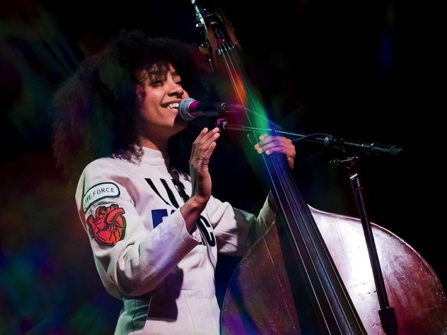 esperanza spalding, wearing a white jumpsuit with a "Life Force" logo, stands next to an upright bass