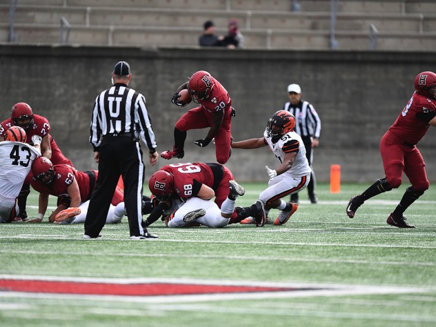 Harvard running back Aaron Shampklin hops over a Harvard teammate with a football in hand.