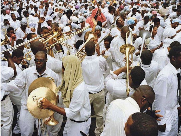 A street crowd of black men and women, all dressed in white, either playing or responding to the playing of dozens of trombones