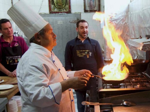 Two men look on while a chef cooks a flaming dish over a stove.