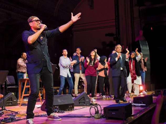 Singer Ali Sethi gestures toward the audience at Sanders Theatre, while students dance on stage behind him.