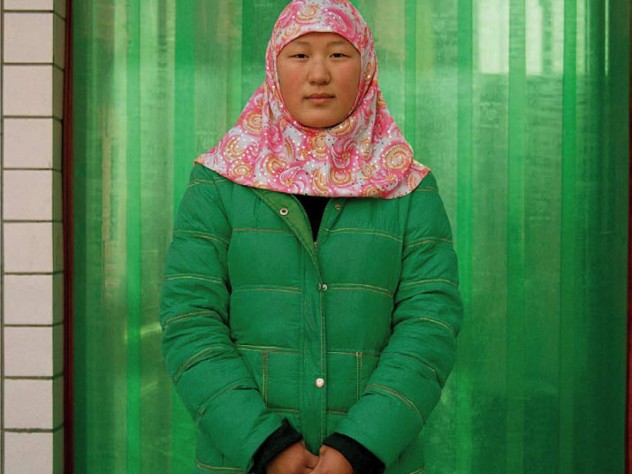 Photograph of a woman in a bright red-and-white hijab and bright green winter coat against a green curtain background, looking directly at the camera