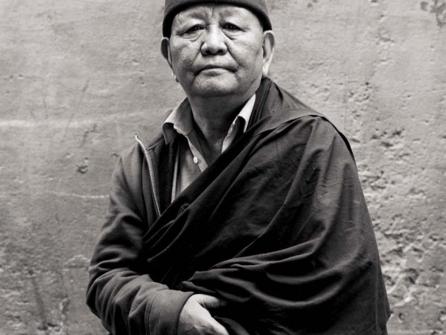 Black-and-white photograph of an older man with a robe draped over one shoulder and a zip-up sweatshirt poking out on the other, and a knit cap with a Nike logo, looks straight at the camera.