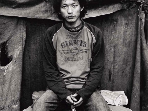Black-and-white photo of a younger man, wearing jeans and an American hockey T-shirt, sitting on a makeshift stone seat, and holding a set of keys, looks straight at the camera.