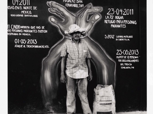 A man in frayed clothing stands in front of a mural marking a timeline of violent acts against immigrants. The mural's dates surround a large pair of hands, handcuffed together with rope.