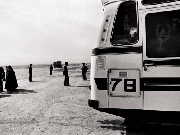 Black-and-white photo of a bus, dominating the right half of the photograph, while a handful of people, and a truck driving toward the horizon, appear as small figures in the left half.