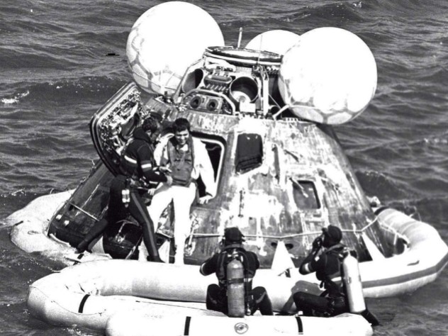 A man in a wetsuit helps an astronaut out of an Apollo capsule floating in the ocean