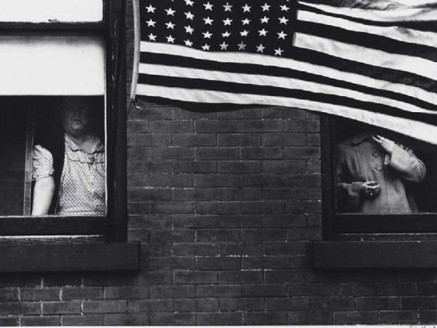 Robert Frank photograph of partially obscured people looking out of windows behind a large American flag