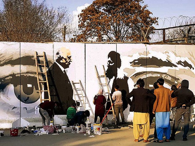 Along a city street, mural on a concrete wall shows two men shaking hands, with a large pair of watchful eyes in the background.