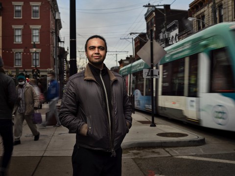 Stephen Gray stands on city sidewalk with street car passing by on right