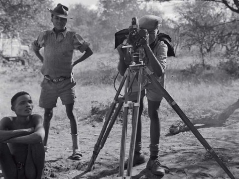 “/Gaishay focusing the camera,” a photograph taken during a 1957-58 ethnographic expedition in Namibia, shows three indigenous boys, one standing behind a camera on a tripod