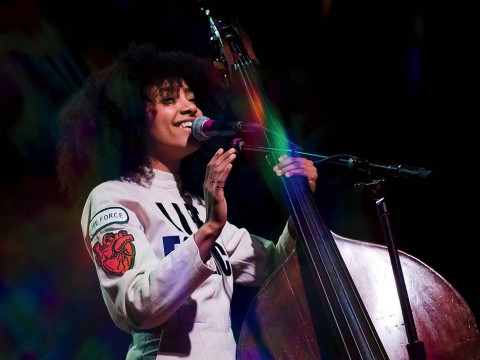 esperanza spalding, wearing a white jumpsuit with a "Life Force" logo, stands next to an upright bass