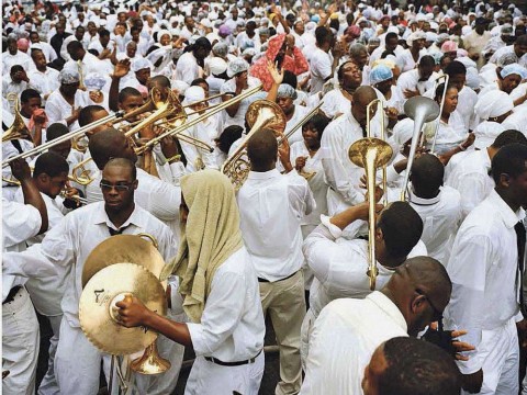 A street crowd of black men and women, all dressed in white, either playing or responding to the playing of dozens of trombones