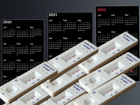 A stack of positive COVID tests in the foreground with 2020-2022 calendars in the background