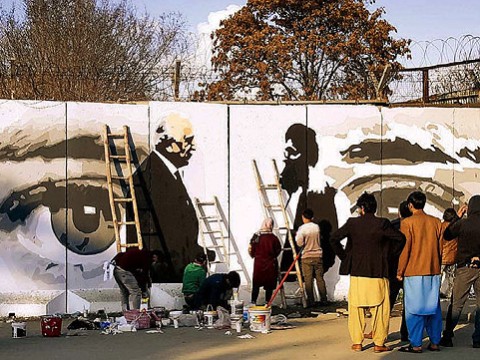 Along a city street, mural on a concrete wall shows two men shaking hands, with a large pair of watchful eyes in  the background.