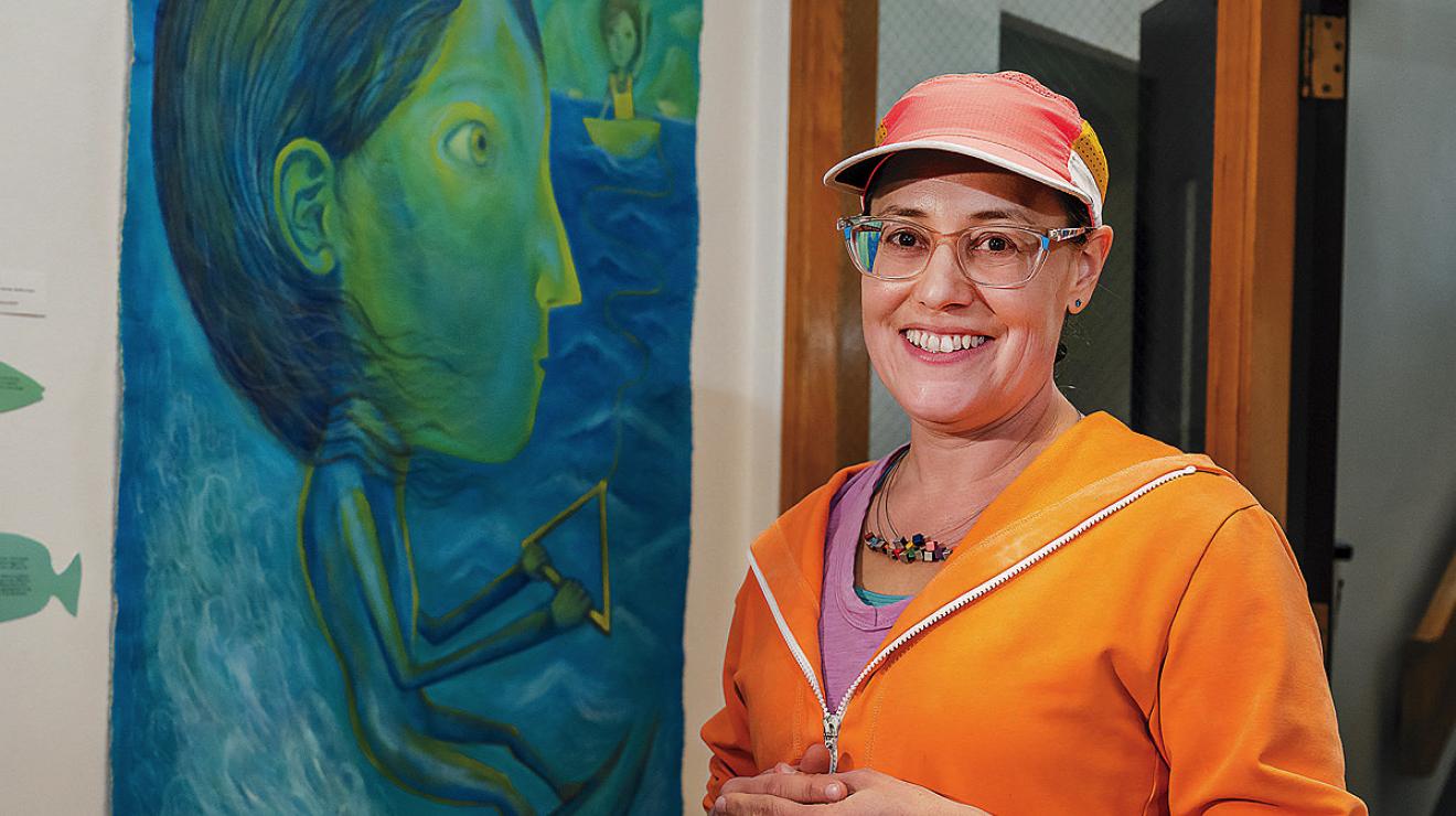 A woman in an orange sweater and cap stands in front of a large blue and green painting of a water-skiing woman.