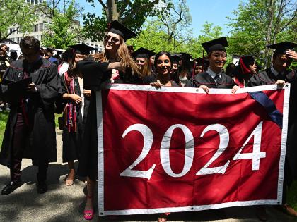 Graduates wearing black caps and gowns processing through Harvard Yard while holding the Class of 2024 banner