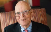 A smiling elderly man in glasses sits comfortably in a striped armchair.